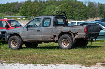 Old pickup truck parked in an overgrown field near a car lot on a sunny day