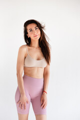 Fototapeta premium Sports woman standing in activewear with abs muscles, looking at camera isolated on white background. Caucasian 30s fitness girl wears beige top, pink leggings. Slim fit smiling brunette, blue eyes.