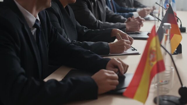 Cropped shot of group of politicians and diplomats in formalwear sitting at meeting table during formal international conference, with hands on folders and clipboards for note taking