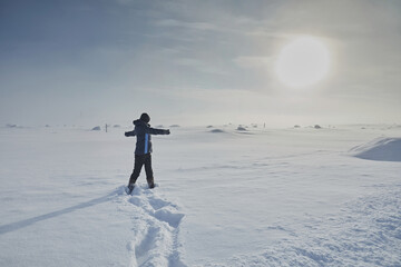 A girl in a blue jacket with outstretched arms against the backdrop of a snowy desert and a sky with the sun. Tundra in the arctic winter © ILIA