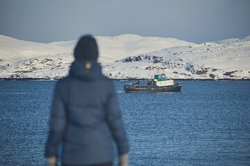 A girl in a blue jacket against the background of a non-freezing sea and a ship in winter, blurry focus, visible on the silhouette