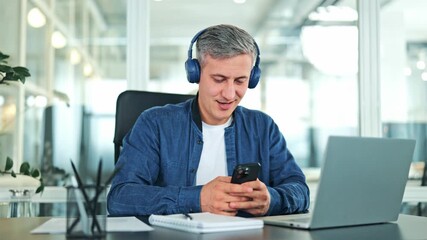 Cheerful male specialist in denim shirt and white t-shirt wearing blue headphones smiling while using smartphone at office desk with laptop.