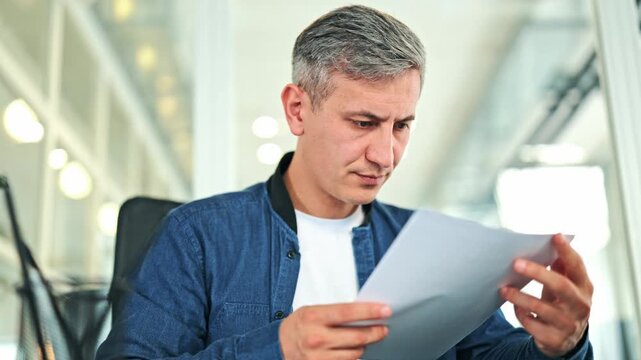 Focused businessman in a blue denim jacket reads critical documents in a bright office. Concerned male evaluates paperwork with a serious expression, reflecting careful consideration of business