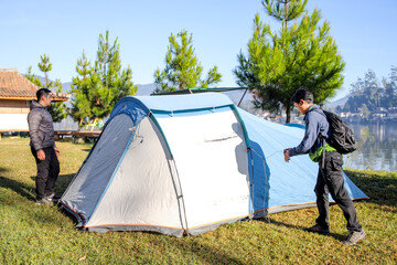 Two Men Securing a Large Blue and White Tent on a Grassy Campsite Near a Tranquil Lake