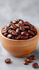 Close up of rich dark roasted coffee beans piled in a rustic wooden bowl with scattered beans on a textured grey surface with soft lighting and a shallow depth of field