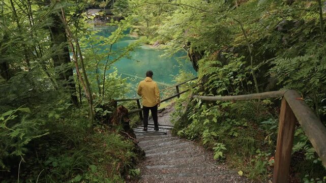 Traveler in a yellow jacket walking down wooden stairs at Blausee in Switzerland, with the turquoise alpine lake view in the background. Scenic summer travel and outdoor exploration in the Swiss Alps