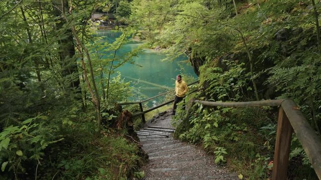 Traveler in a yellow jacket climbing wooden stairs at Blausee in Switzerland, with the turquoise alpine lake view in the background. Summer adventure travel lifestyle in nature.