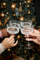 Multiethnic group of young adult hands holding crystal glasses making toast in front of decorated Christmas tree celebrating festive holiday together