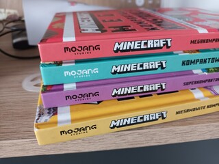 Naklejka premium A stack of colorful official Minecraft handbooks by Mojang Studios in the Finnish language, resting on a wooden shelf