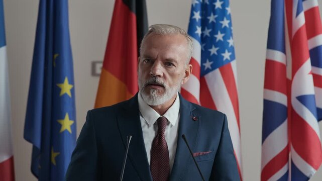 Chest up of confident Caucasian male speaker delivering official speech at global conference podium surrounded by flags of different nations symbolizing cooperation and politics