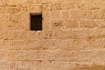 Malta - A rustic stone wall, crafted from unevenly cut blocks, features a small, dark window protected by metal bars, showcasing ancient architectural style.