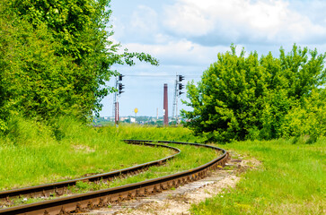 Curved railway tracks passing through green nature towards industrial city horizon. Railway tracks bend through lush greenery toward city skyline, symbolizing travel, transition, connection between