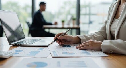 Businesswoman analyzes charts with laptop in office setting coworker visible in background.