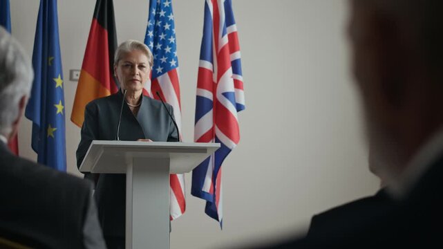 Confident mature Caucasian woman in elegant formalwear giving speech at podium with flags of different countries in background symbolizing diplomacy, leadership, and global cooperation