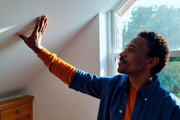 Black young man touching sloped ceiling with right hand while standing near window, looking upward with slight smile, sunlight illuminating face and hand, indoor setting