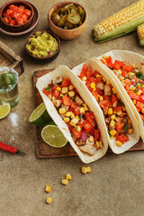 Tacos with chicken, tomatoes and corn with guacamole and salsa sauce, lime, fresh corn cobs, chili peppers and tequila shots on beige concrete background. Mexican party snacks