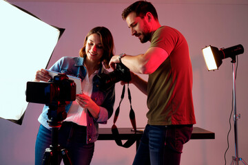 Young woman and young man collaborating in photography studio, adjusting camera settings and reviewing images on digital camera with professional lighting equipment