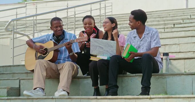 Indian young adult gen z lady woman male man group sitting on stair hold guitar play music sing song have fun joy. Happy pupil girl boy group spend break time relax mood clap hands enjoy new say place