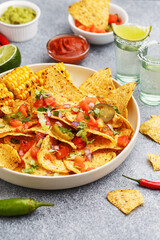 Tequila shots and nachos corn chips served with chopped tomatoes, onion and greens, marinated jalapeno, grilled corn and various sauces on gray concrete background. Mexican party snacks