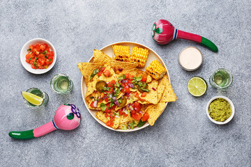 Nachos corn chips served with chopped tomatoes, onion, greens and grilled corn, marinated jalapeno, guacamole and lime on gray concrete background with tequila shots and maracas. Overhead view