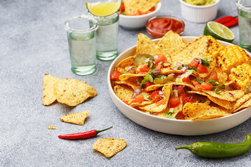 Tequila shots and nachos corn chips served with chopped tomatoes, onion and greens, marinated jalapeno, grilled corn and various sauces on gray concrete background. Mexican party snacks