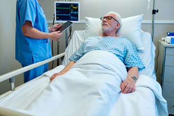 Senior man lying in hospital bed wearing medical gown, looking at young male nurse standing beside bed holding digital tablet and monitoring vital signs