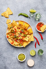 Nachos corn chips served with chopped tomatoes, onion, greens and grilled corn, marinated jalapeno, guacamole and lime on gray concrete background with tequila shots. Overhead view, flat lay
