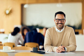 Fototapeta premium Smiling Man in Glasses at Table in Modern Café Office Setting, With Background Team Meeting