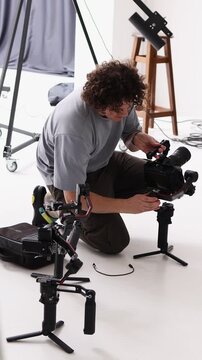 A professional videographer adjusts an electronic gimbal stabilizer and a cinema camera in a studio setting. Focus on expertise, technology, and filmmaking preparation.
