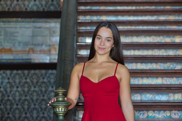 Young woman, brunette, Latina, wearing a red dress, looking at the camera, happy and smiling, leaning on a staircase railing in the Spain square in Seville. Concept: youth, travel, destinations.