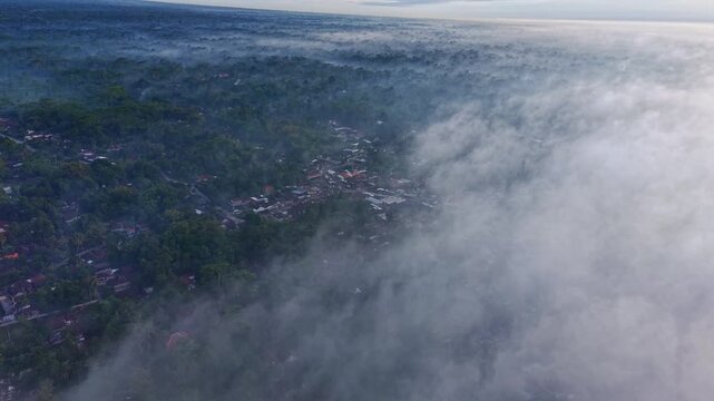 Aerial view of the landscape, where mist hangs low over the forest and buildings are visible, Klaten Regency, Central Java, Indonesia.