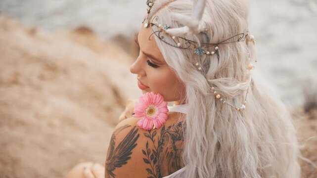 fantasy portrait young elf woman holding pink gerbera flower. close-up beauty face sexy eyes lips adult fairy girl, tiara diadem on head blonde ash hair. myth faun, pixie lady sits on riverbank sea