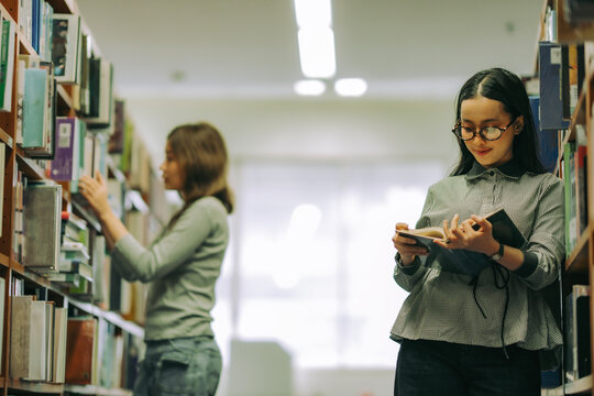 Woman finding academic book in university library, depicting education, research skills, and pursuit of lifelong learning. Woman with glassess leaning against book shelf concentrate for reading book.
