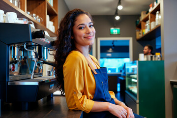 Portrait of young Hispanic woman smiling while standing behind counter in coffee shop, wearing apron, long curly hair visible, espresso machine and shelves in background