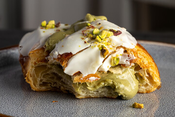 Primer plano de un croissant relleno de crema de pistacho, cubierto con glaseado blanco y trozos de pistacho. Imagen detallada que resalta la textura crujiente del hojaldre y la cremosidad del relleno