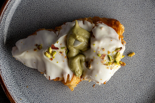 Primer plano de un croissant relleno de crema de pistacho, cubierto con glaseado blanco y trozos de pistacho. Imagen detallada que resalta la textura crujiente del hojaldre y la cremosidad del relleno