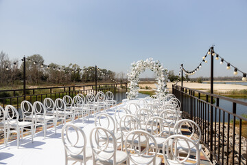 A wedding ceremony with white chairs, a path and an arch of white flowers on the lake shore 