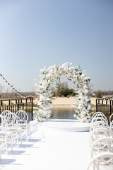 A wedding ceremony with white chairs, a path and an arch of white flowers on the lake shore 