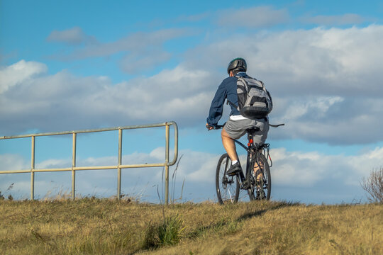 Calgary, Alberta, Canada. Oct 28, 2025. A lone cyclist wearing a helmet and backpack, riding a bicycle up a grassy hill against a bright blue sky with clouds.