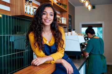 Portrait of young woman with long curly hair smiling and leaning on counter in modern coffee shop while female coworker preparing drinks at espresso machine in background