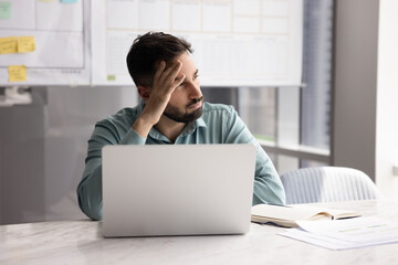 Pensive young businessman sitting at desk looking aside from notebook