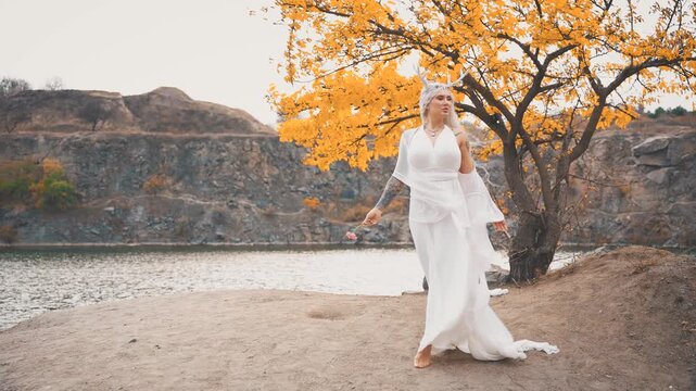 fantasy young elf woman beauty face walking by river bank water sea, autumn nature yellow tree. sexy adult fairy girl fashion model long blonde ash hair white dress fly in wind. myth faun pixie lady