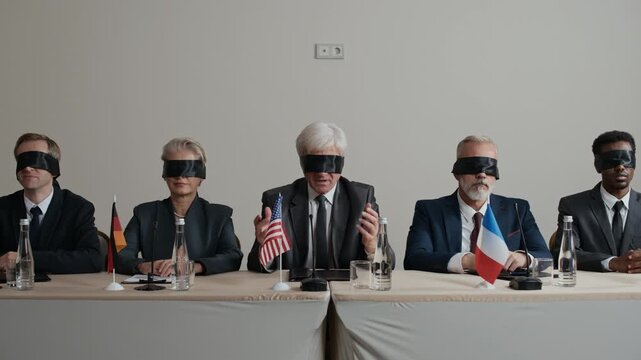 Dolly-in shot of senior politician gesturing his hands while delivering speech during press conference, sitting at table with other political country representatives, all wearing black blindfolds