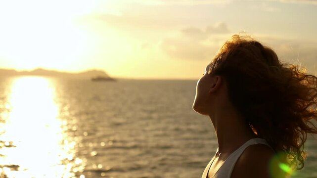 Silhouette of a happy woman enjoying the fresh air on a sea cruise, her curly hair fluttering in the wind against a beautiful golden sunset, feeling relaxed, free and enjoying her vacation