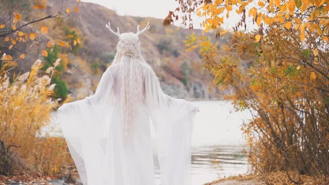 fantasy young elf woman beauty face walking by river bank water sea, autumn nature yellow tree. sexy adult fairy girl fashion model long blonde ash hair white dress fly in wind. myth faun pixie lady