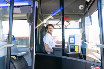 Smiling male bus driver sitting at the steering wheel in his cabin, providing public transport service