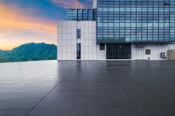 Empty square floor and modern office building with green mountain landscape at sunset.
