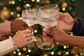 Diverse group of young adult hands clinking crystal glasses in celebratory toast in front of decorated Christmas tree, showing multiethnic friendship and holiday gathering