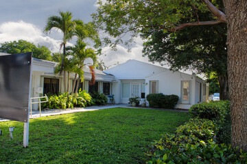 A white tropical-style house surrounded by lush greenery and palm trees under a partly cloudy sky. The well-maintained lawn and modern architecture create a serene suburban atmosphere.