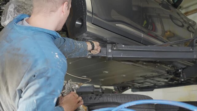 Man on garage floor inserts tube attached to spray gun into car door sill cavity to apply lanolin rustproofing treatment, caps opening when finished